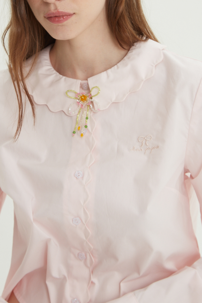 Camisa rosa de poplin de largo hasta la cadera con terminación en forma de onditas en cuello, puños y cartera y botones a tono en forma de flor. 