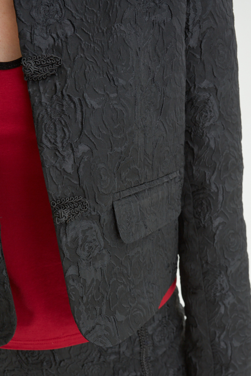 Saco negro de brocato con textura de rosas y alamares de pasamanería, mangas princesas y cuello mao, con bolsillos con solapa delanteros y largo a la cadera. 