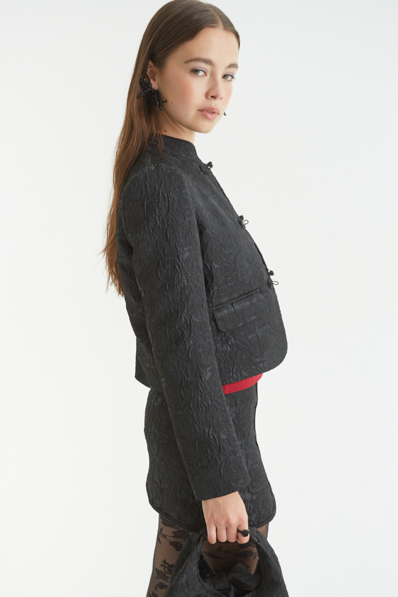 Saco negro de brocato con textura de rosas y alamares de pasamanería, mangas princesas y cuello mao, con bolsillos con solapa delanteros y largo a la cadera. 