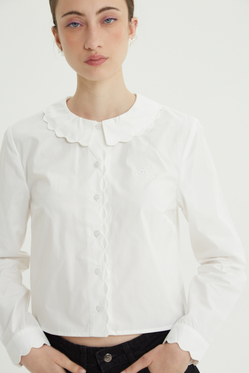 Camisa blanco de poplin de largo hasta la cadera con terminación en forma de onditas en cuello, puños y cartera y botones a tono en forma de flor. 