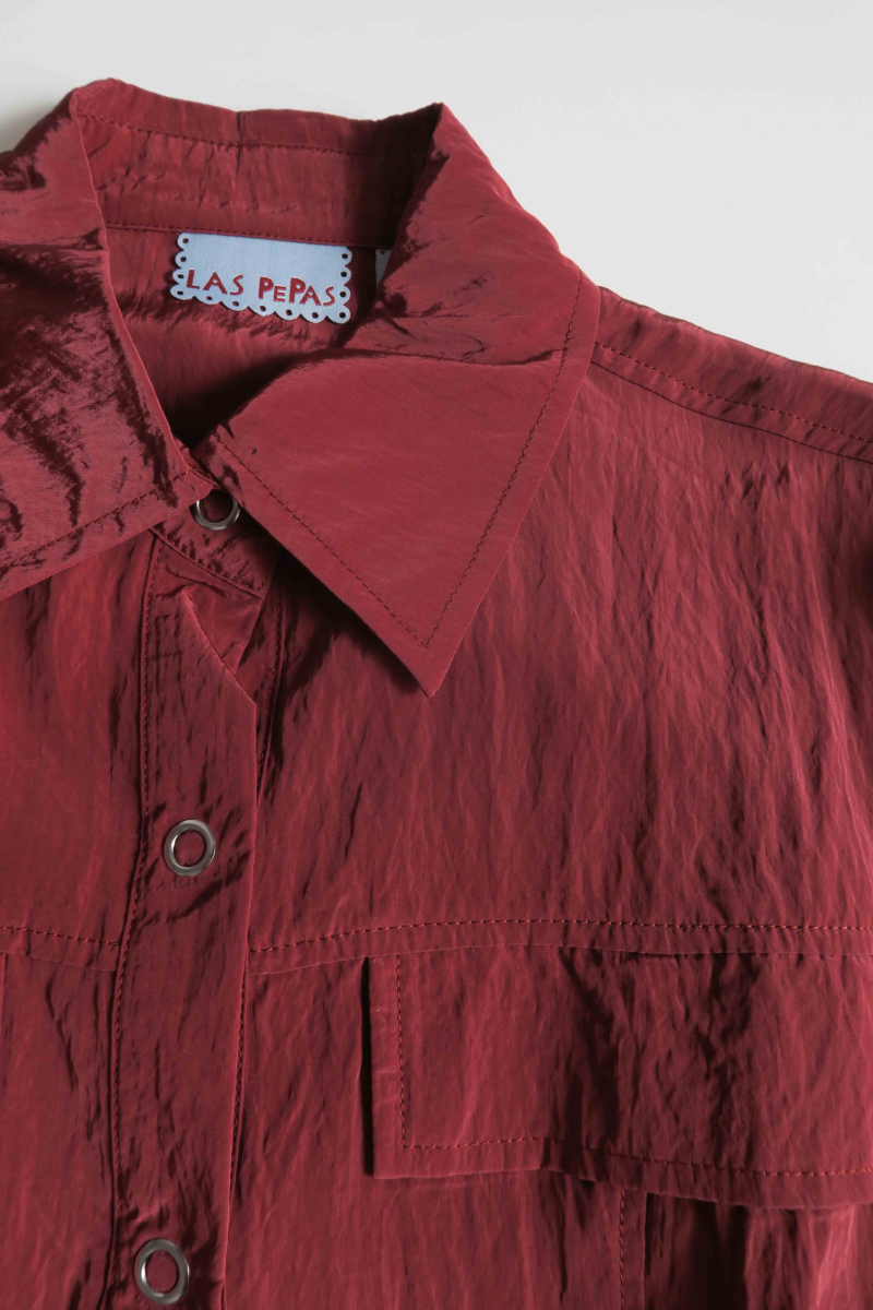 Camisa bordo manga larga con textura arrugada y brillo sutil, bolsillos con solapa y cinturones en la cintura. 