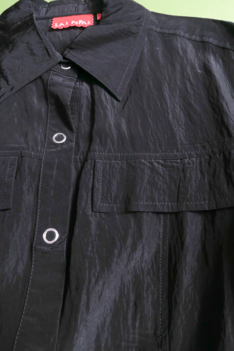 Camisa negra manga larga con textura arrugada y brillo sutil, bolsillos con solapa y cinturones en la cintura. 