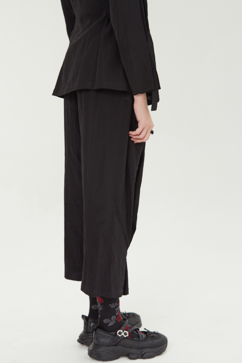 Pantalón negro con textura arrugada y brillo sutil, con pinza en la pierna ajustada al tobillo, cintura con cinturón y bolsillos traseros.
