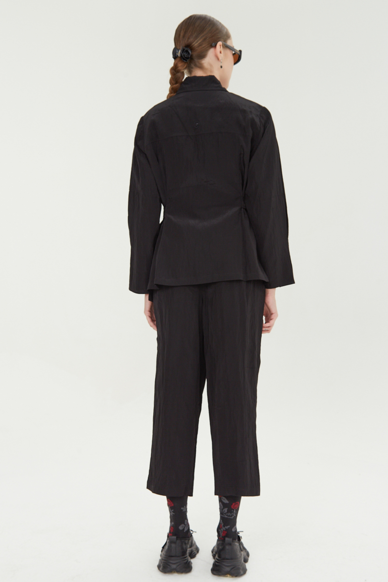 Pantalón negro con textura arrugada y brillo sutil, con pinza en la pierna ajustada al tobillo, cintura con cinturón y bolsillos traseros.