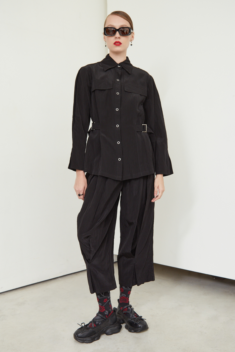Pantalón negro con textura arrugada y brillo sutil, con pinza en la pierna ajustada al tobillo, cintura con cinturón y bolsillos traseros.
