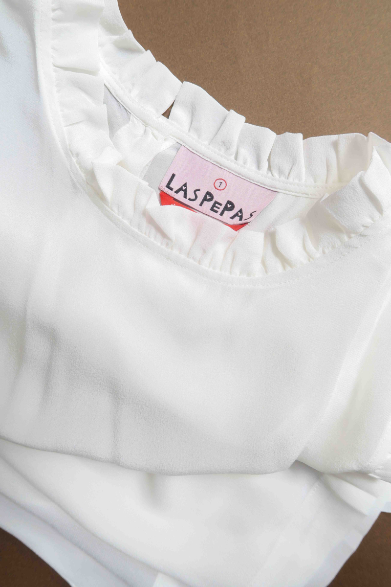 Musculosa cruda de seda con volados en cuello y sisas, de corte recto y larga a la cadera con cuello mao y botones en la espalda. 
