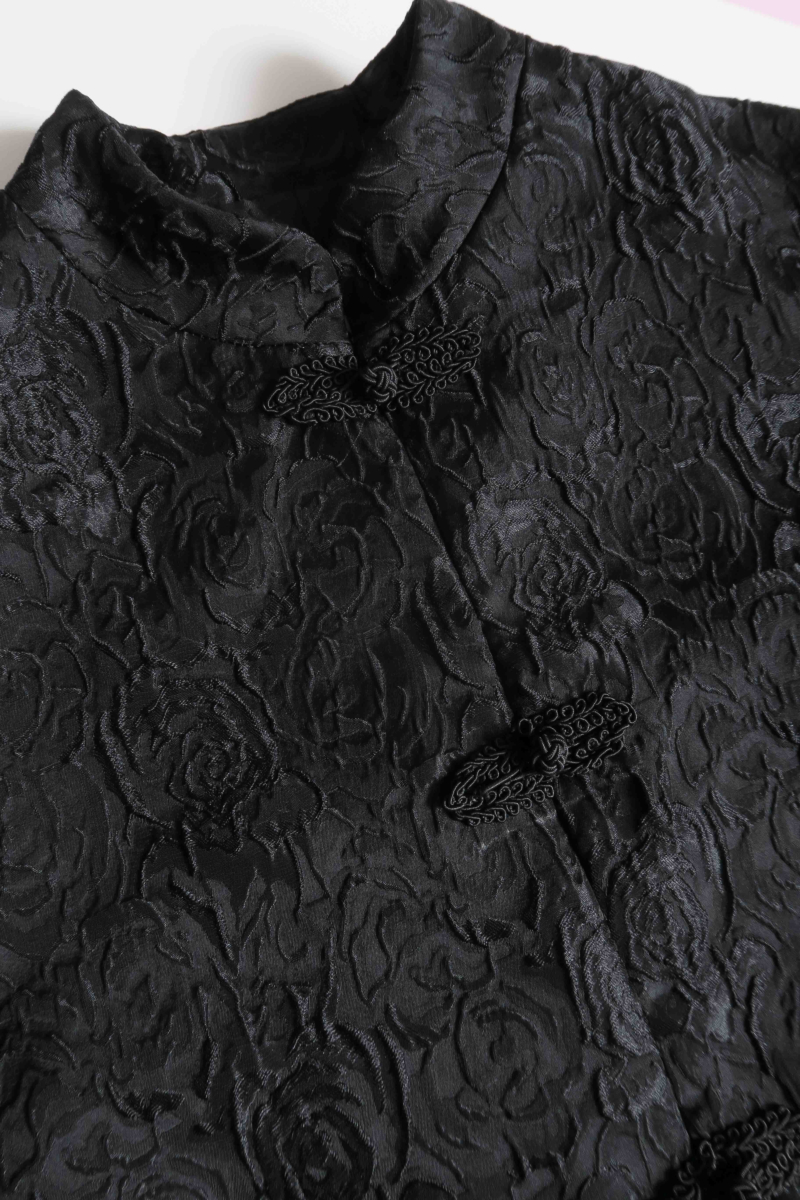 Saco negro de brocato con textura de rosas y alamares de pasamanería, mangas princesas y cuello mao, con bolsillos con solapa delanteros y largo a la cadera. 